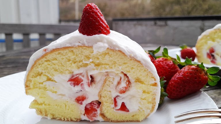 ein Stück Erdbeer-Sahne-Biskuitrolle auf einem weißen Teller, daneben ganze Erdbeeren und eine Kuchengabel
