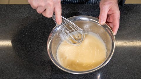 Leckeres Rezept für Ananas-Kokos-Kuchen: Ein Ei wird in einer silbernen Schüssel mit dem Schneebesen aufgeschlagen.