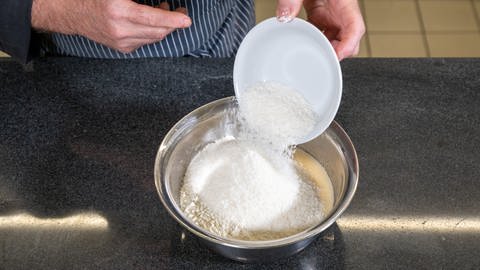 Leckeres Rezept für Ananas-Kokos-Kuchen: Die Mehl- und Triebmittel-Mischung sowie die Kokosraspeln werden in einer silbernen Schüssel in die Joghurtmasse gegeben und untergemischt.