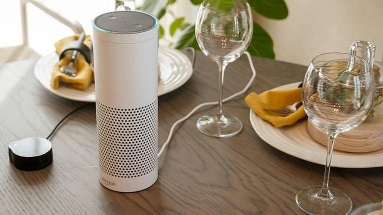 Alexa & Co. - so funktionieren Smart Speaker und Sprachassistent. Ein Smart Speaker steht neben Gläsern und Tellern auf einem gedecktem Tisch.