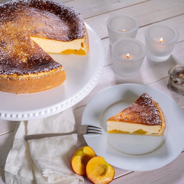 Ein Käsekuchen mit Pfirsich auf einer Kuchenplatte, ein Stück davor angeschnitten auf einem Teller: Bei diesem Rezept verbindet sich eine leckere Füllung mit Quark und ein feiner Boden aus Mürbeteig. (Foto: SWR, Corinna Holzer)