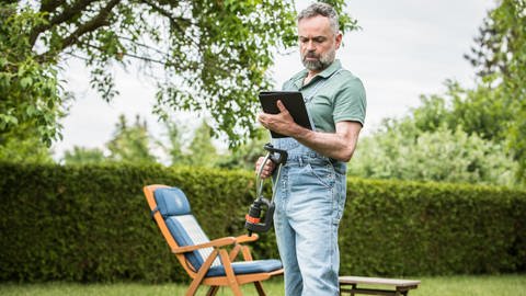 Ein Mann mit einem Rasensprinkler in der Hand schaut im Garten in ein Tablet: Mit Technik kann es sich der Faule leicht machen bei der Pflege der Pflanzen.