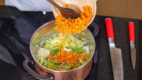 Gewürfelte Karotten für die Lauchcremesuppe werden in einen Topf gegeben. Später wird die Suppe mit Croutons und Speck serviert.