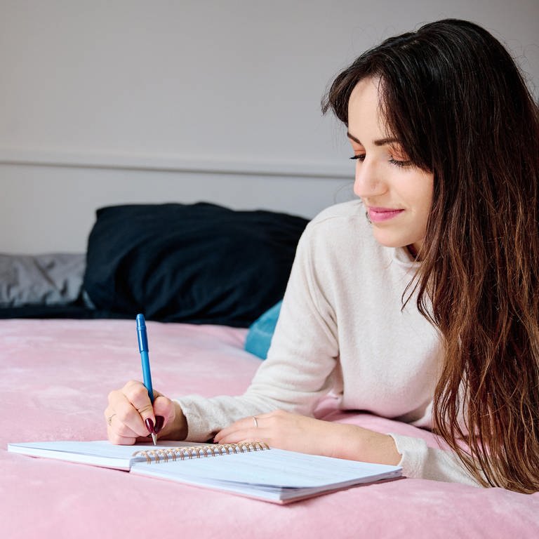 Dankbarkeitstagebuch: Eine Frau mit langen dunklen Haaren liegt im Bett und schreibt in ein leeres Buch mit einem blauen Stift. Die positiven Gedanken dabei scheinen sie zum Lächeln zu bewegen und ihrer Stimmung gut zu tun.