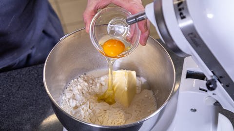Ein Mann schüttet aus einer Schale ein aufgeschlagenes Ei in eine Rührschüssel mit Mehl und Butter: Der Hefeteig für Apfeltaschen geligt einfach mit zimmerwarmen Zutaten.