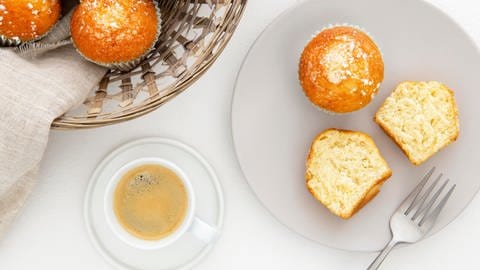 Auf einem Teller sind zwei Zitronenmuffins, einer von beiden ist halbiert. Daneben stehen eine Tasse Kaffee und ein Korb mit weiteren Muffins.