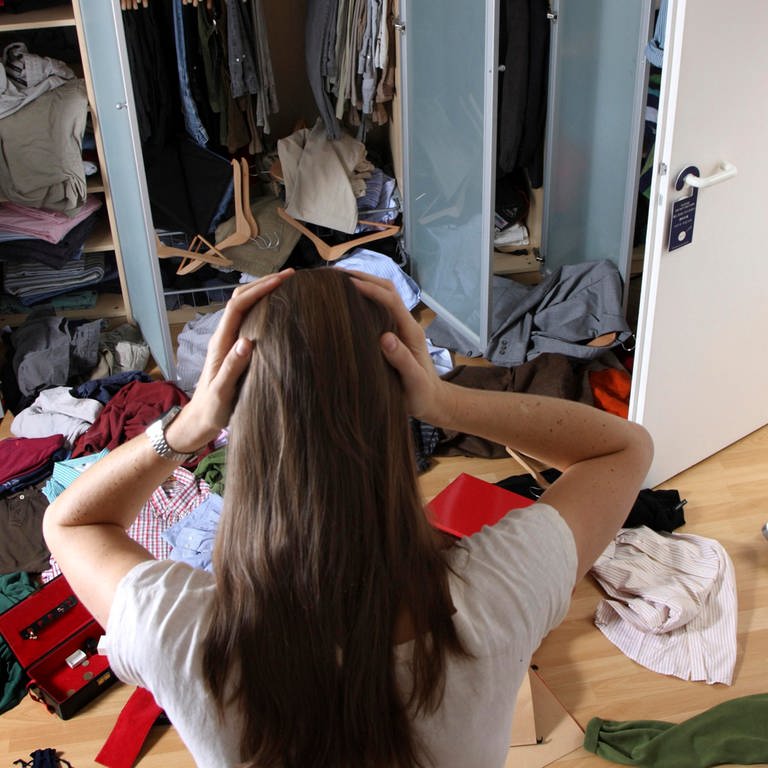 Einbruch – was tun? Eine junge Frau von hinten gesehen steht nach einem Einbruch in einer verwüsteten Wohnung und greift sich mit beiden Händen an den Kopf.
