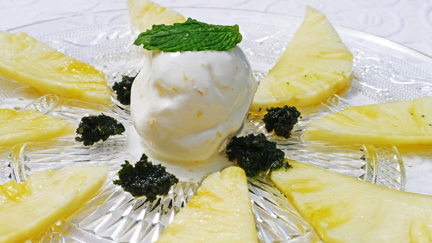 Rezept für ein Dessert: Zitroneneis mit in Rum marinierter Ananas auf einem Glasteller. Auf der Eiskugel in der Mitte liegt ein Minzblatt. (Foto: SWR, Stefanie Kühn)