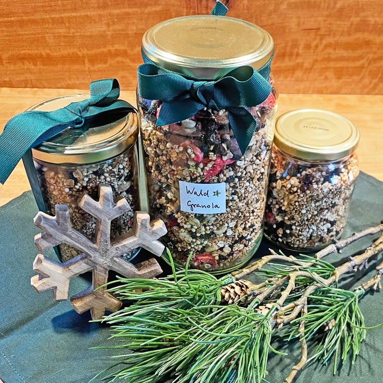 Rezept für Knuspermüsli (Granola): Drei Gläser mit Müsli gefüllt, mit Schleifen dekoriert auf einem Tisch, grüne Zweige und ein Holzstern als Deko daneben.