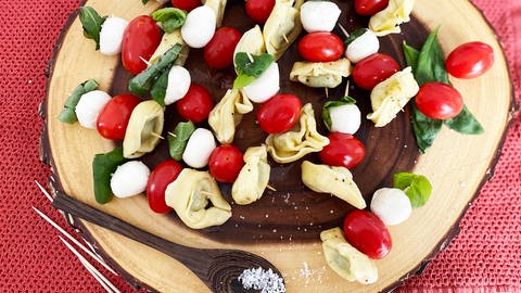Rezept für Tortellini-Spieße: Spieße mit Tortellini, Tomaten, Basilikum auf einer Holzplatte. Daneben ein Holzlöffel mit grobem Salz (Foto: SWR, Sarah Korz)