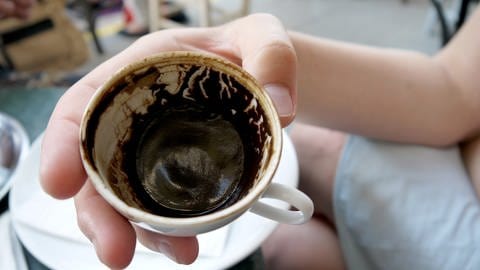 Silvester Kaffeesatz lesen anstelle von Bleigießen -- Eine Hand hält eine weiße Kaffeetasse mit Kaffeesatzspuren über einer Untertasse (Foto: dpa Bildfunk, Picture Alliance)