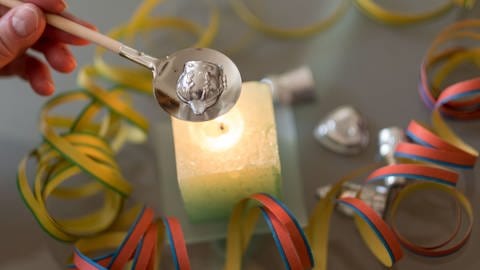Bleigießen an Silvester über Kerze - eine Hand hält einen Löffel mit Holzstil, auf dem ein Stück Blei in Form einer Krone zum Schmelzen liegt, darunter ist eine angezündete quadratische Kerze umgeben von Luftschlangen (Foto: dpa Bildfunk, Picture Alliance)