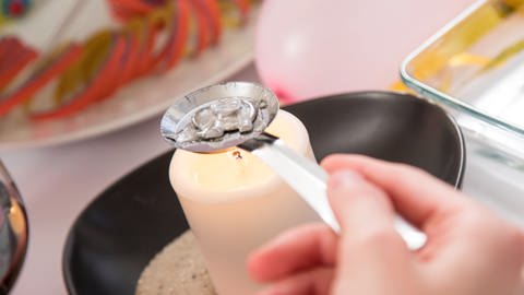 Zinngießen an Silvester - eine Frauenhand hält einen Löffel mit Holzstiel wie beim Bleigießen mit einem Stück Zinn darin, das am Schmelzen ist über eine flammenden runden Kerze, daneben steht eine recheckig Glasschüssel mit Wasser (Foto: dpa Bildfunk, Picture Alliance)