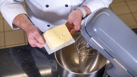 Zwei Hände halten eine offene Packung Butter über einer Küchennmaschine, die Butter soll für das Gebäck Flammendes Herz aufgeschlagen werden (Foto: SWR)