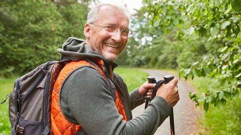 Rente und Altersvorsorge - entspannt in den Ruhestand: Ein Senior geht in der grünen Natur wandern. Er hält Nordic Walking-Stöcke in der Hand, hat eine orange Weste an und trägt einen Rucksack auf dem Rücken. Er dreht sich zur Kamera und lächelt.