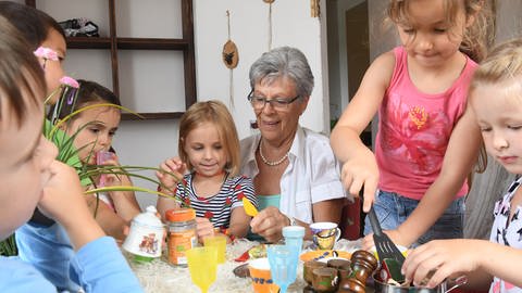 Rente und Altersvorsorge - entspannt in den Ruhestand: Eine Rentnerin sitzt in einer Kindertagesstätte mit mehreren Kindern an einem Tisch und lächelt.