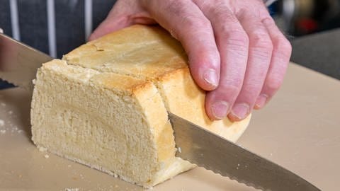 Eine Hand hält eine Hälfte von dem fertig gebackenen Zwieback fest und die andere schneidet mit einem Brotmesser Scheiben ab.