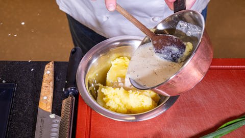 Eine weiße Flüssigkeit mit schwarzen Punkten wird mithilfe eines Kochlöffels in eine Schüssel mit einer gelben Masse geschüttet - dem Kartoffelpüree.