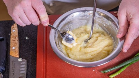 Ein Stück Butter auf einem Löffel wird in eine gelbe, cremige Massein einer Metallschüssel, dem Kartoffelpüree gegeben. In der Schüssel steckt ein großer Löffel.