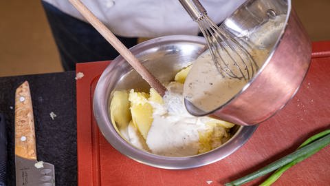 Eine weiße Flüssigkeit mit schwarzen Punkten wird mithilfe eines Schneebesens in eine Schüssel mit einer gelben Masse geschüttet, in der sich auch ein Kochlöffel befindet - dem Kartoffelpüree.