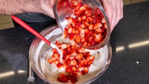 Aus einer Metallschüssel werden klein geschnittene Erdbeeren in eine andere Metallschüssel mit der weißen Quark-Creme für die frische Erdbeertorte mit Biskuitboden geschüttet. 