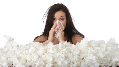 Eine Frau sitzt in Taschentüchern. Heuschnupfen und Allergien lösen häufig Schnupfen aus.