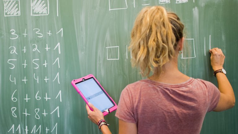 Digitalpakt: Lehrer und Schüler sollen digitale Medien künftig stärker nutzen, das Bild zeigt eine Lehrerin mit einem tablet in der Hand, die an einer Tafel Matheformeln schreibt (Foto: dpa Bildfunk, Picture Alliance)