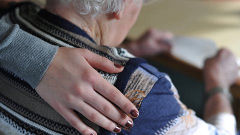 Eine junge Frau hat ihre Hand auf der Schulter einer älten Frau gelegt, die in einem Seniorenheim am Tisch sitzt.