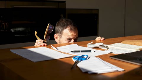 Besorgter Mann versteckt sich vor Rechnungen auf Schreibtisch (Foto: IMAGO, Westend61)