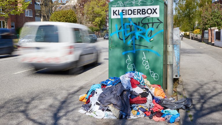 Ein Kleiderberg liegt vor einem Altkleidercontainer mit der Aufschrift «Kleiderbox» am Straßenrand (Foto: dpa Bildfunk, picture alliance/dpa | Georg Wendt)