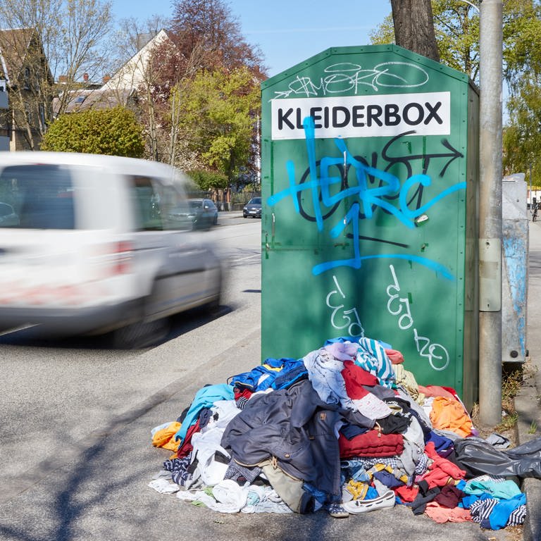 Ein Kleiderberg liegt vor einem Altkleidercontainer mit der Aufschrift «Kleiderbox» am Straßenrand (Foto: dpa Bildfunk, picture alliance/dpa | Georg Wendt)