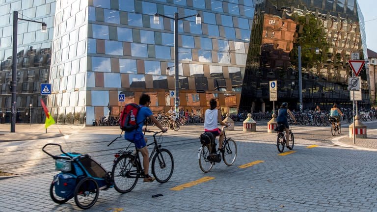 Vor der Universitätsbibliothel in Freiburg fahren mehrere Fahrradfahrer mit sommerlicher Kleidung vorbei