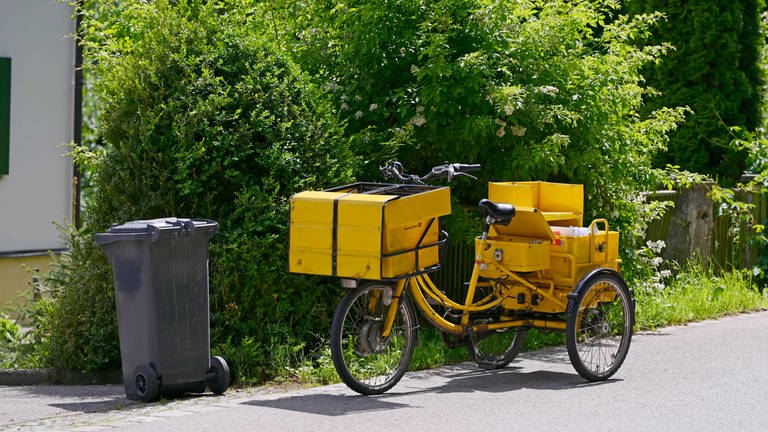 Ein Briefträger der Deutschen Post hat sein Transportrad vor einem Haus abgestellt um die Post an die Haustüre zu bringen.