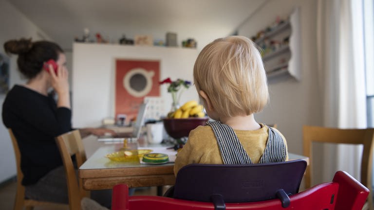 Eine Frau sitzt im Homeoffice an ihrem Laptop und telefoniert, während ihr Kind neben ihr in einem Kinderstuhl am Tisch sitzt. (Foto: dpa Bildfunk, picture alliance/Christian Beutler/KEYSTONE/dpa)