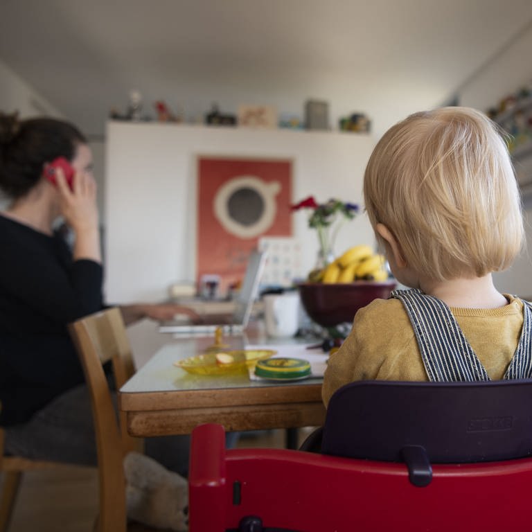 Eine Frau sitzt im Homeoffice an ihrem Laptop und telefoniert, während ihr Kind neben ihr in einem Kinderstuhl am Tisch sitzt. (Foto: dpa Bildfunk, picture alliance/Christian Beutler/KEYSTONE/dpa)