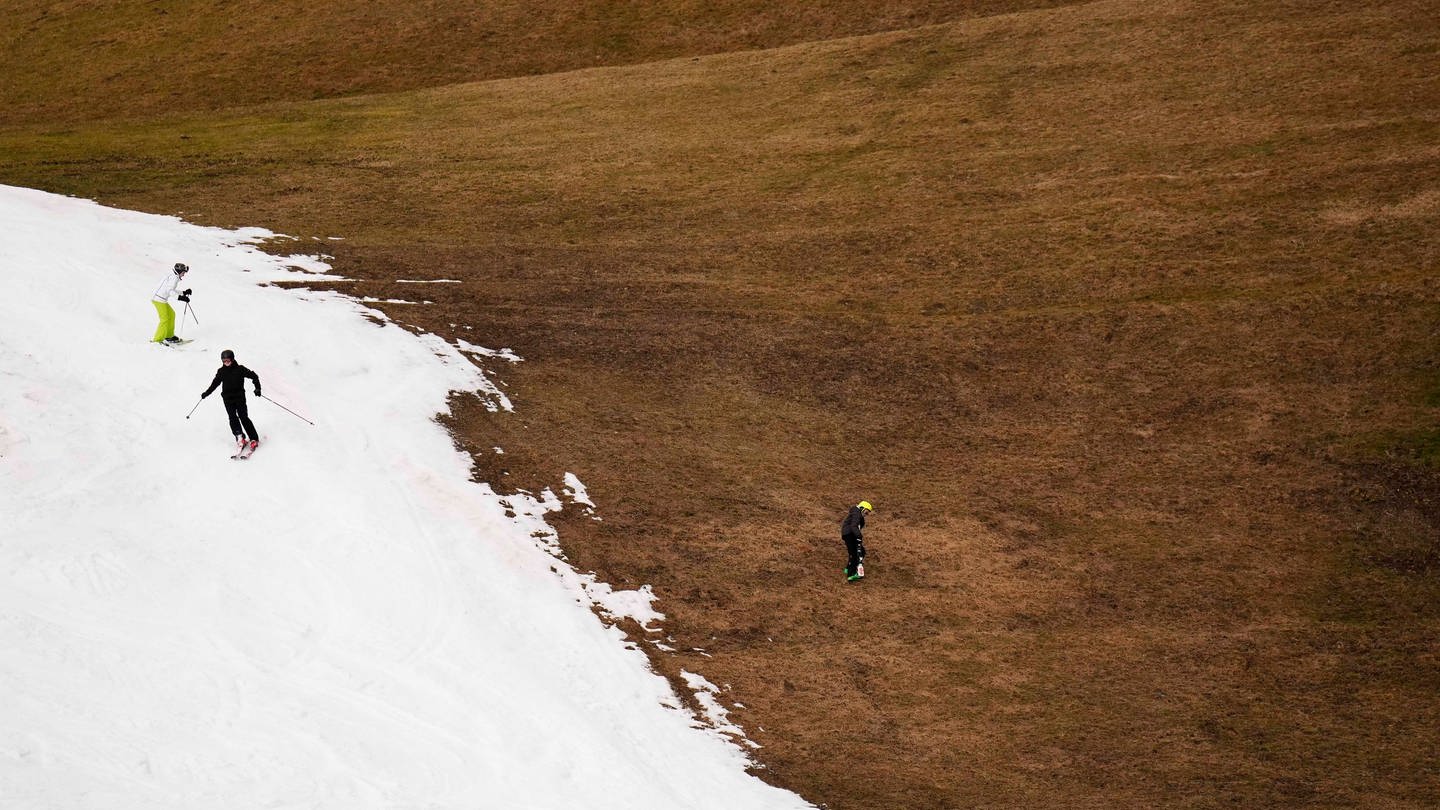 Skifahrer fahren eine Piste neben einer Wiese, auf der kein Schnee liegt, herab. (Foto: picture-alliance / Reportdienste, dpa Bildfunk, picture alliance/dpa/AP | Matthias Schrader)