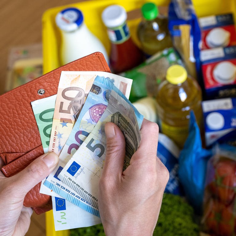 Eine Einkaufskiste mit Lebensmitteln steht auf einem Küchentisch, während eine Frau Euro-Banknoten in den Händen hält. 