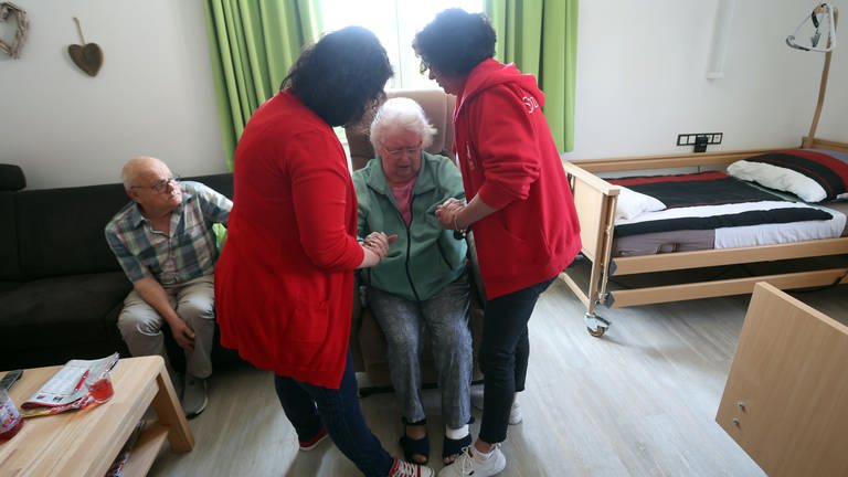 Pflegerinnen helfen pflegebedürftiger Frau aus einem Sessel