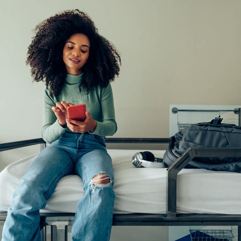 Das Handy als Schlüssel für Hiotelzimmer. Junge Frau sitzt mit einem Smartphone auf einem Hotelbett.  (Foto: Adobe Stock, AdobeStock/Carlo)