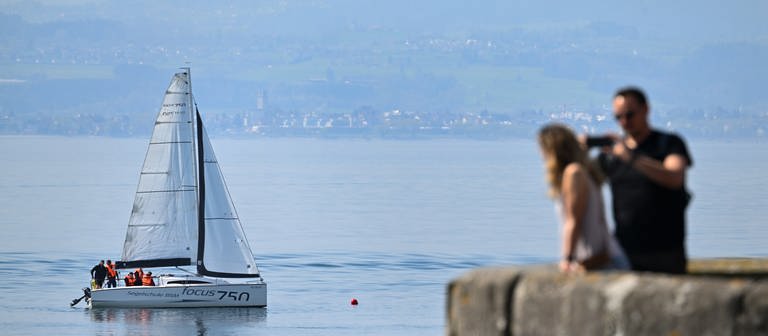 Ein Segelboot einer Segelschule trainieren vor Kressbronn auf dem Bodensee, während ein Paar im Vordergrund auf der Terasse des Schlosses Montfort steht. Der See ist ruhig, es geht kaum Wind. (Foto: dpa Bildfunk, picture alliance/dpa | Felix Kästle)