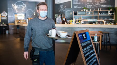 Ein Kellner läuft mit Mundschutz und einem Tablett durch ein Café. (Foto: dpa Bildfunk, picture alliance/dpa | Julian Stratenschulte)