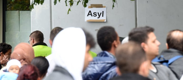  In der Landeserstaufnahmestelle (LEA) für Flüchtlinge in Karlsruhe (Baden-Württemberg) warten am 08.10.2014 Flüchtlinge auf ihre Registrierung. Im Hintergrund ist ein Papier an der Wand angebracht, auf dem das Wort "Asyl" steht.  (Foto: dpa Bildfunk, picture alliance / dpa | Uli Deck / Archivbild)
