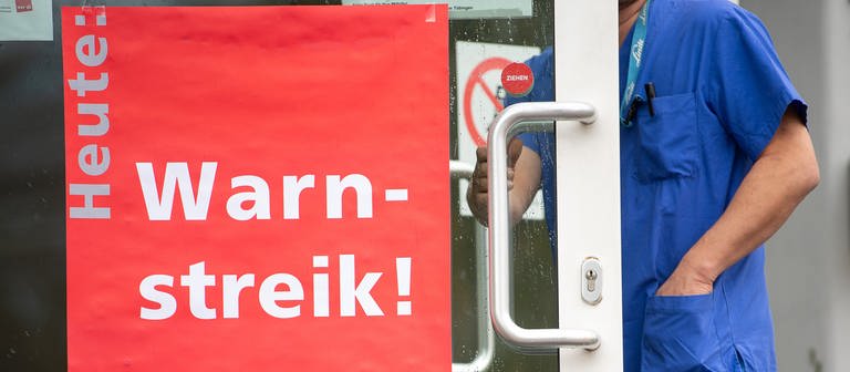 Ein Mitarbeiter des Universitätsklinikums Tübingen geht während eines Warnstreiks an den Uni-Kliniken durch eine Tür mit einem Plakat, auf dem "Heute: Warnstreik!" steht. (Foto: dpa Bildfunk, picture alliance/dpa | Sebastian Gollnow (Archivbild))