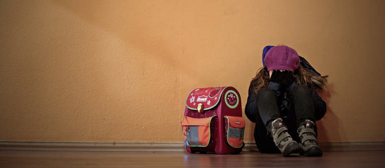 Ein Mädchen sitzt neben ihrem Schulranzen auf dem Boden und hält die Hände vor das Gesicht. (Foto: dpa Bildfunk, picture alliance/dpa | Nicolas Armer)