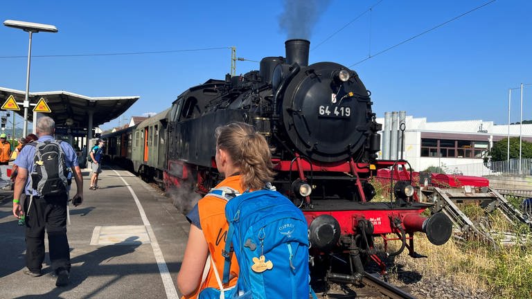 Eine Wanderin mit Rucksack steht auf dem Bahnsteig vor der schwarzen Lok der schwäbischen Waldbahn. (Foto: SWR)