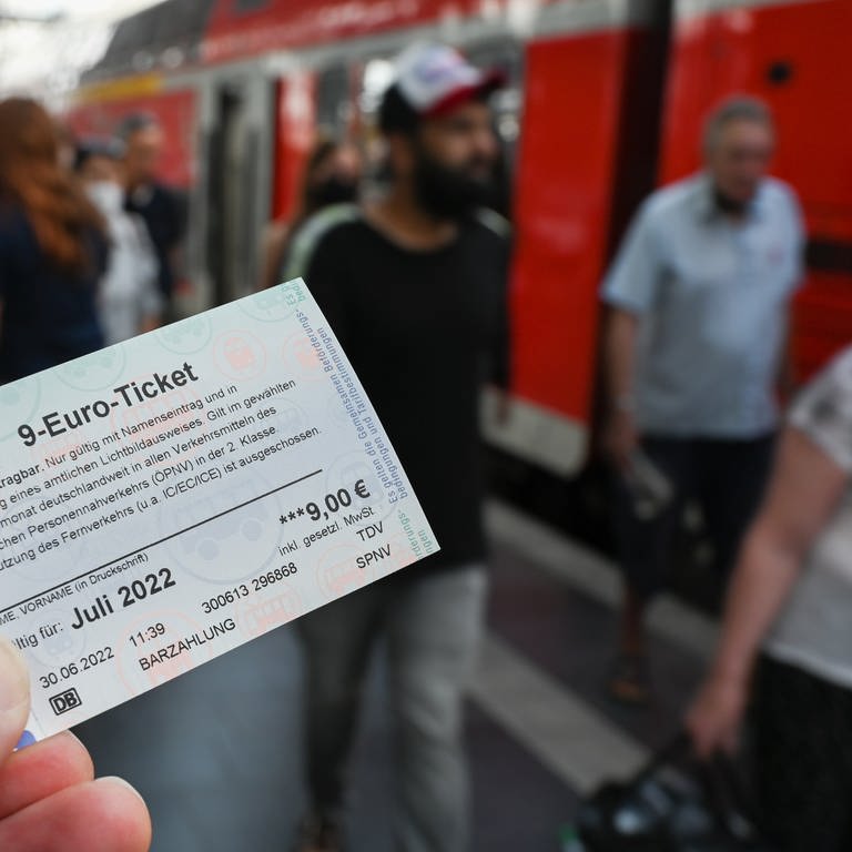 Symbolbild: Ein 9-Euro-Ticket für Juli 2022 wird am Hauptbahnhof hochgehalten, während Zugreisende aus einer Regionalbahn aussteigen. (Foto: dpa Bildfunk, picture alliance/dpa | Arne Dedert)