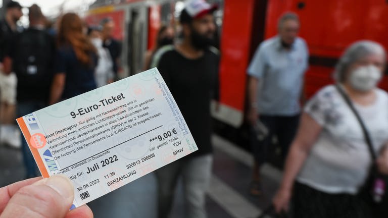 Symbolbild: Ein 9-Euro-Ticket für Juli 2022 wird am Hauptbahnhof hochgehalten, während Zugreisende aus einer Regionalbahn aussteigen. (Foto: dpa Bildfunk, picture alliance/dpa | Arne Dedert)