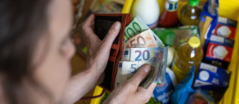 Eine Frau hält Geldscheine in einer Geldbörse über einer Einkaufskiste mit Lebensmitteln, der auf einem Küchentisch steht. (Foto: dpa Bildfunk, picture alliance/dpa | Hendrik Schmidt)