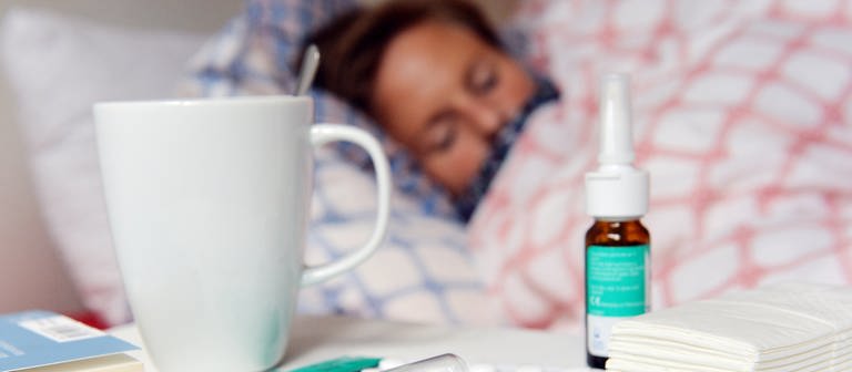 Eine Frau liegt mit Grippe im Bett, auf dem Nachtisch Fieberthermometer, Teetasse, Taschentücher, Tabletten und Nasenspray (Foto: dpa Bildfunk, Maurizio Gambarini)