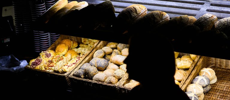 picture alliancedpa | Julian Stratenschulte (Foto: dpa Bildfunk, Eine Bäckereifachverkäuferin steht am frühen Morgen in der bewusst sparsam beleuchteten Bäckerei)
